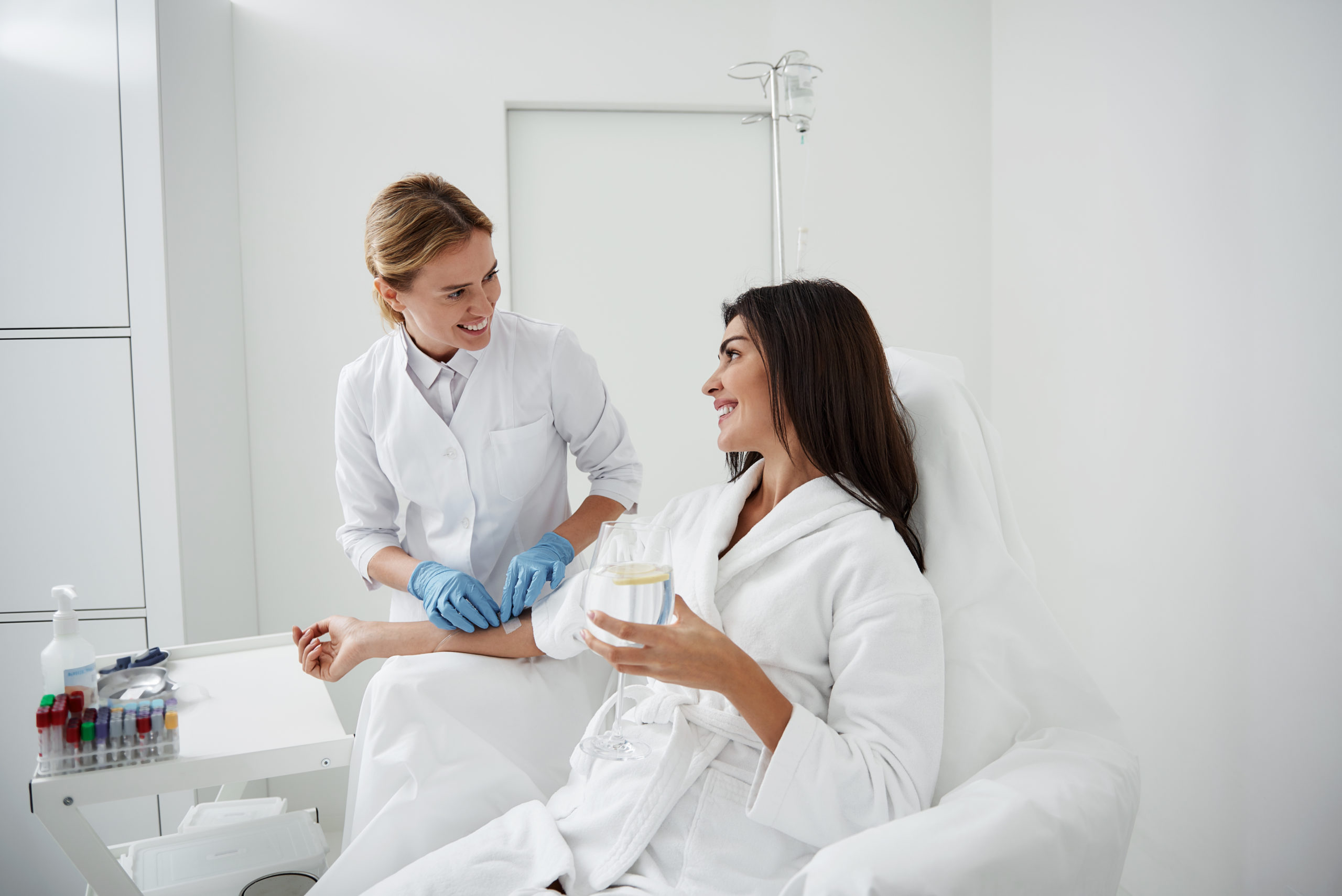 Portrait of charming woman sitting in armchair and holding glass of lemon water while doctor in sterile gloves checking IV infusion